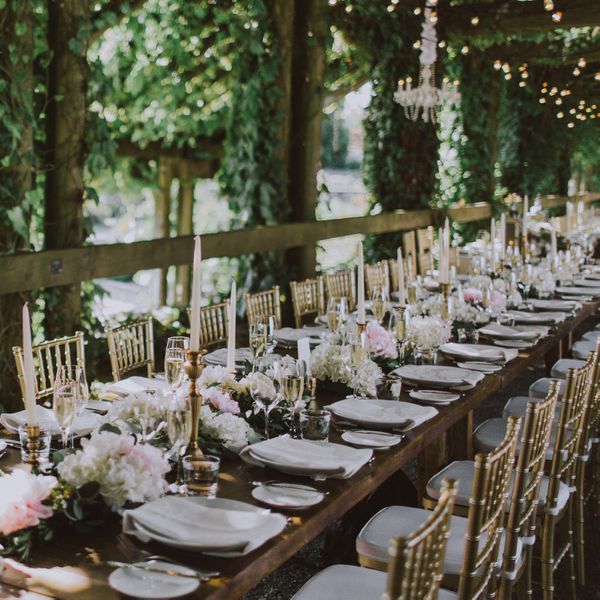 A wedding reception table with greenery, hanging lights, pink flowers, and gold chairs.